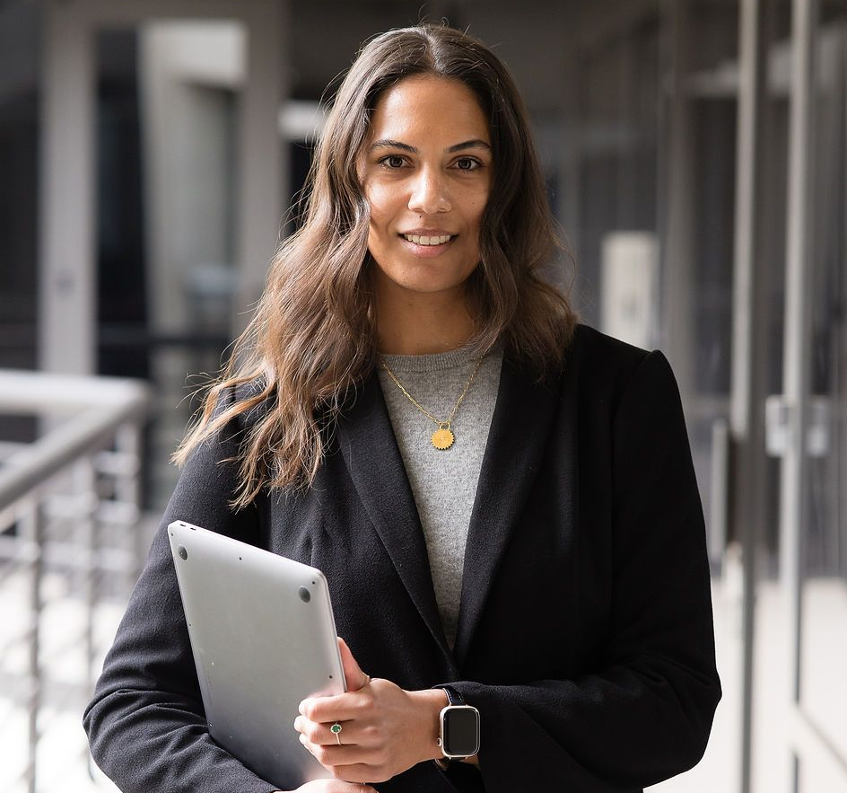 A smiling professional wearing a black blazer and gray top holds a tablet while standing in a modern hallway.