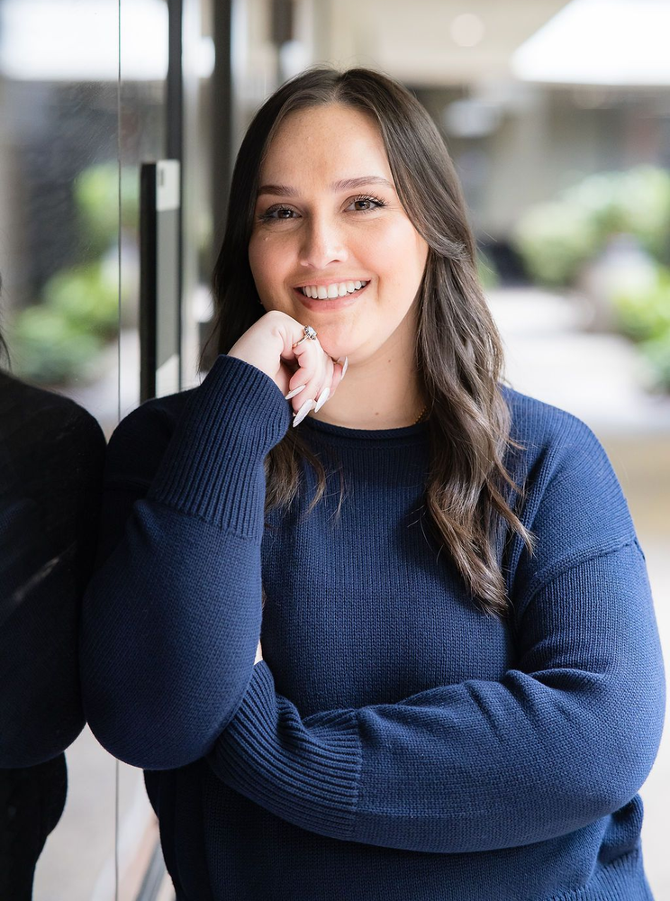 A smiling person wearing a dark blue sweater, leaning against a glass wall in an outdoor setting.