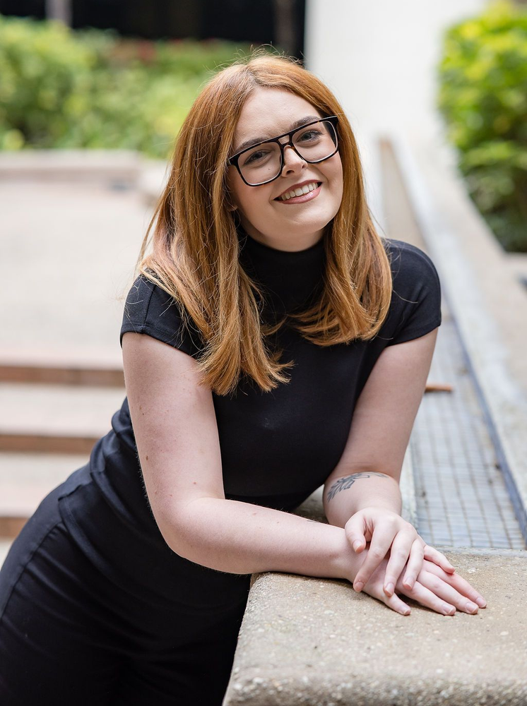 A person with auburn hair and glasses leans on a stone ledge outdoors, smiling at the camera.