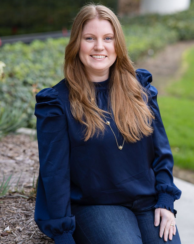 A smiling person with long light-brown hair wearing a dark blue puff-sleeve top outdoors.