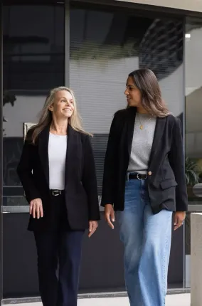Two people wearing black blazers walk together and talk in front of a modern glass building.