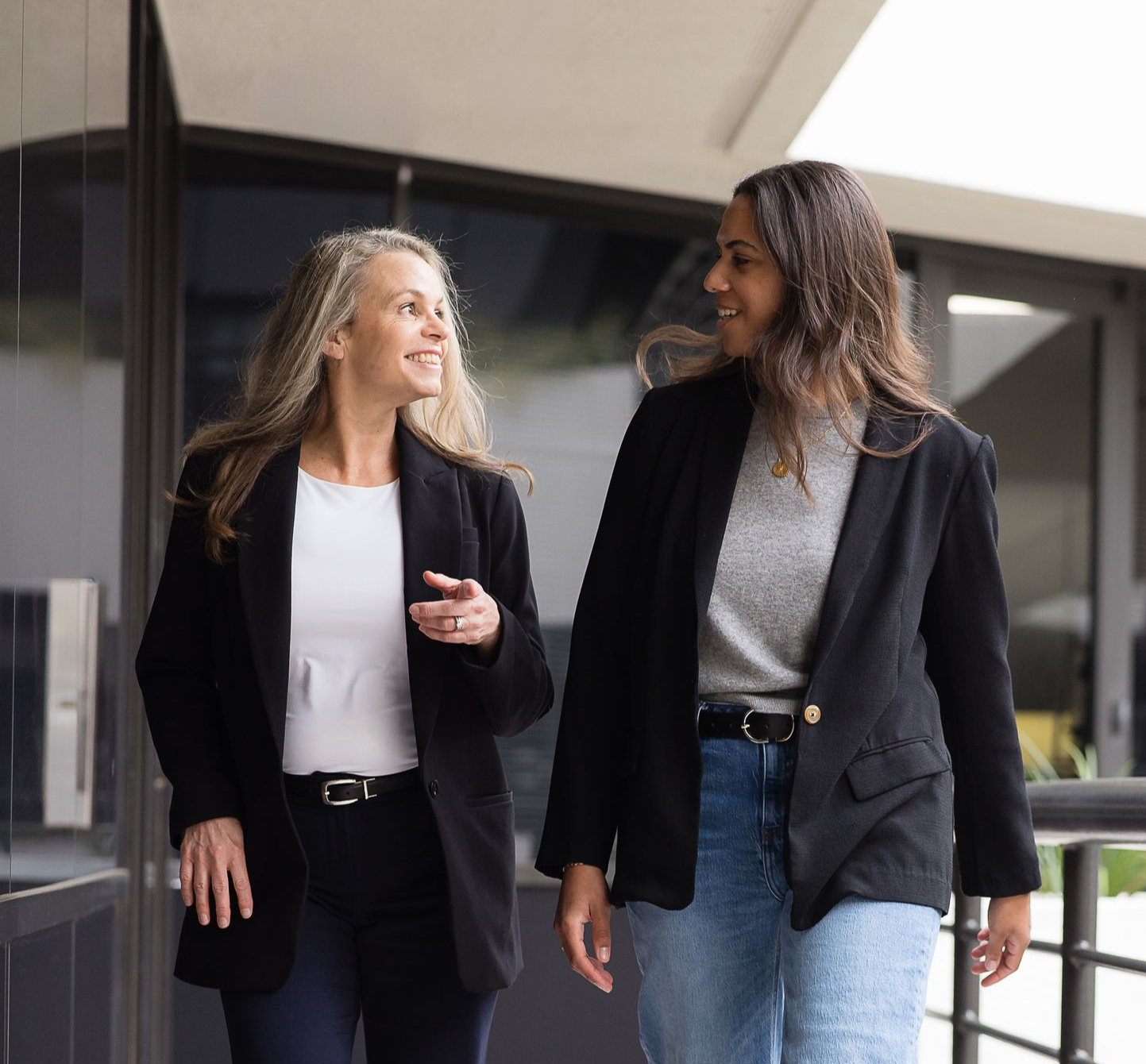 Two people walk outdoors, wearing black blazers and smiling while talking to each other near a glass-paneled building.