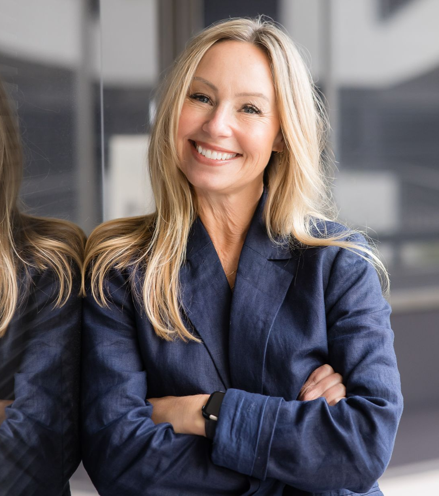 A smiling person with long blonde hair, wearing a navy blue blazer, stands with arms crossed against a glass background.
