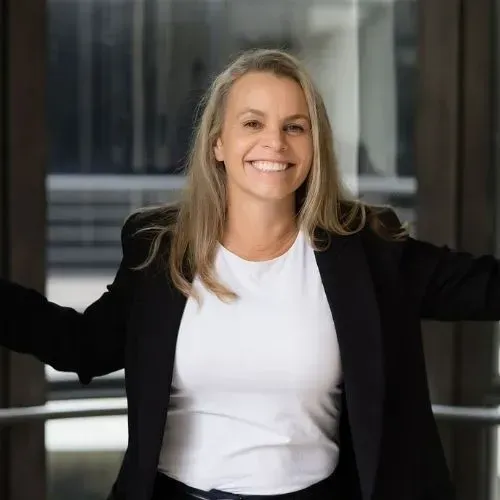 A smiling professional in a white shirt and black blazer stands in front of a windowed office setting.