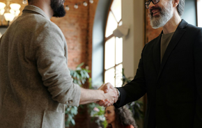 Two professionals in blazers shaking hands in a bright, modern office with a blurred person visible in the background.