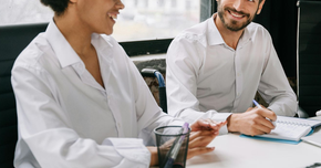 Two professionals in white shirts sit at a table, smiling and collaborating while reviewing notes in an office setting.