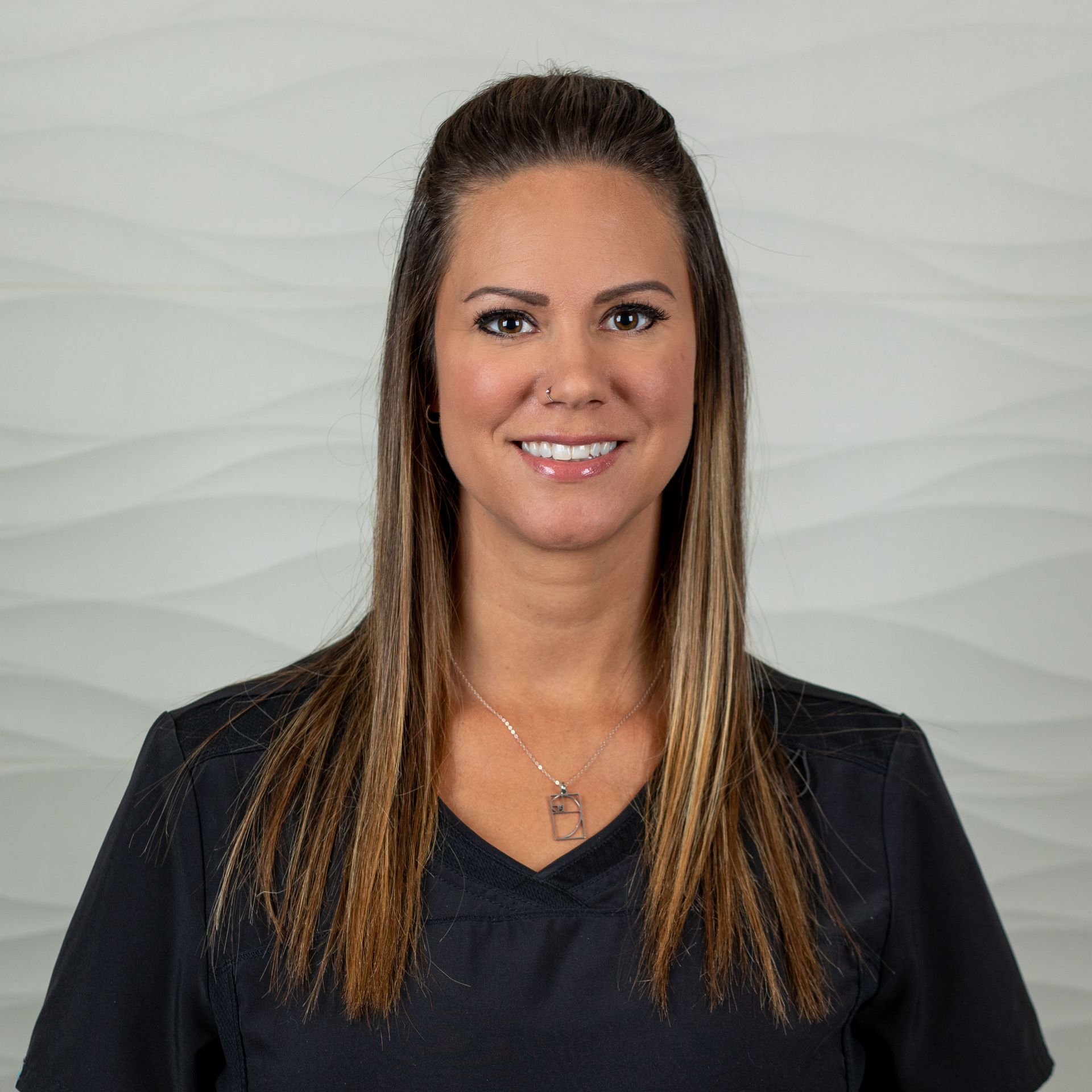 A person with long, highlighted brown hair wearing a dark scrub top, smiling against a neutral, wavy-patterned background.