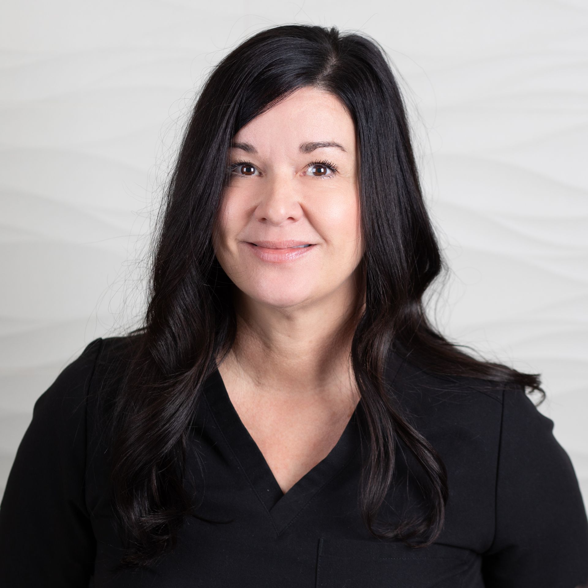 A person with long dark hair, wearing a black top, looks toward the camera against a plain, textured white background.