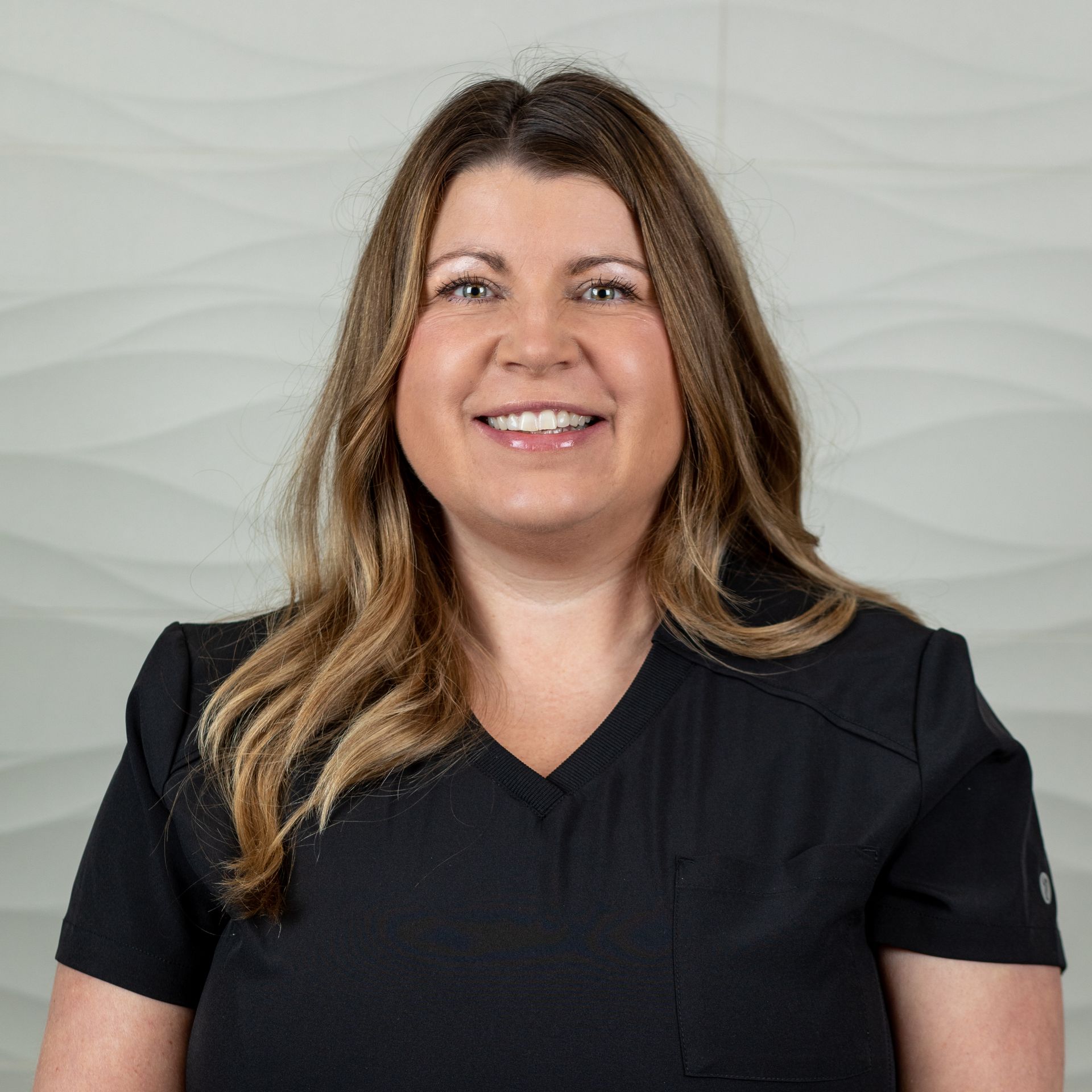 Smiling person with shoulder-length wavy hair wearing a black V-neck scrub top against a textured light gray wall.