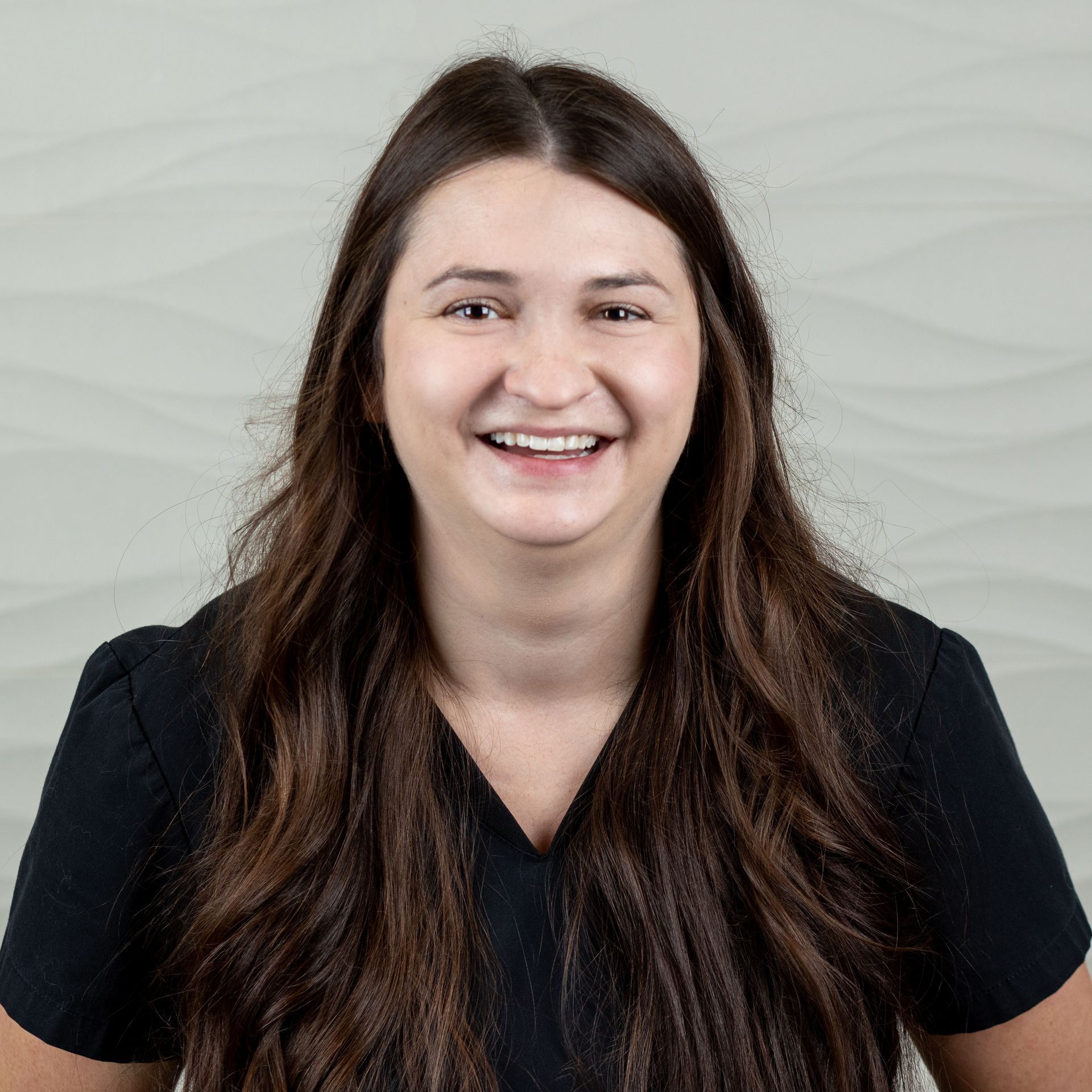 A smiling person with long brown hair wearing a black V-neck top against a light gray, wavy textured background.