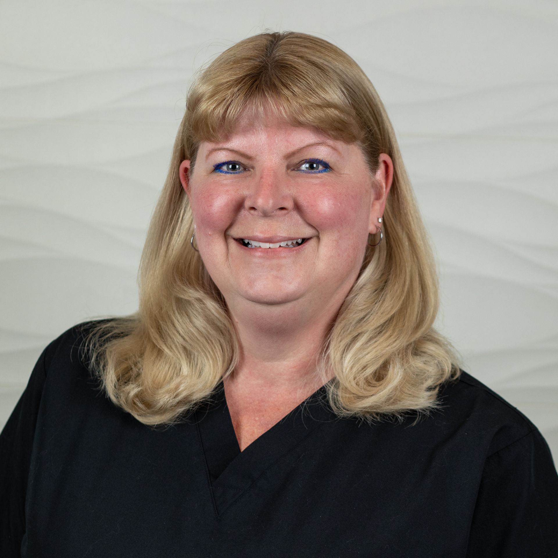 A smiling blonde person wearing a black scrub top, posed against a light gray textured background.