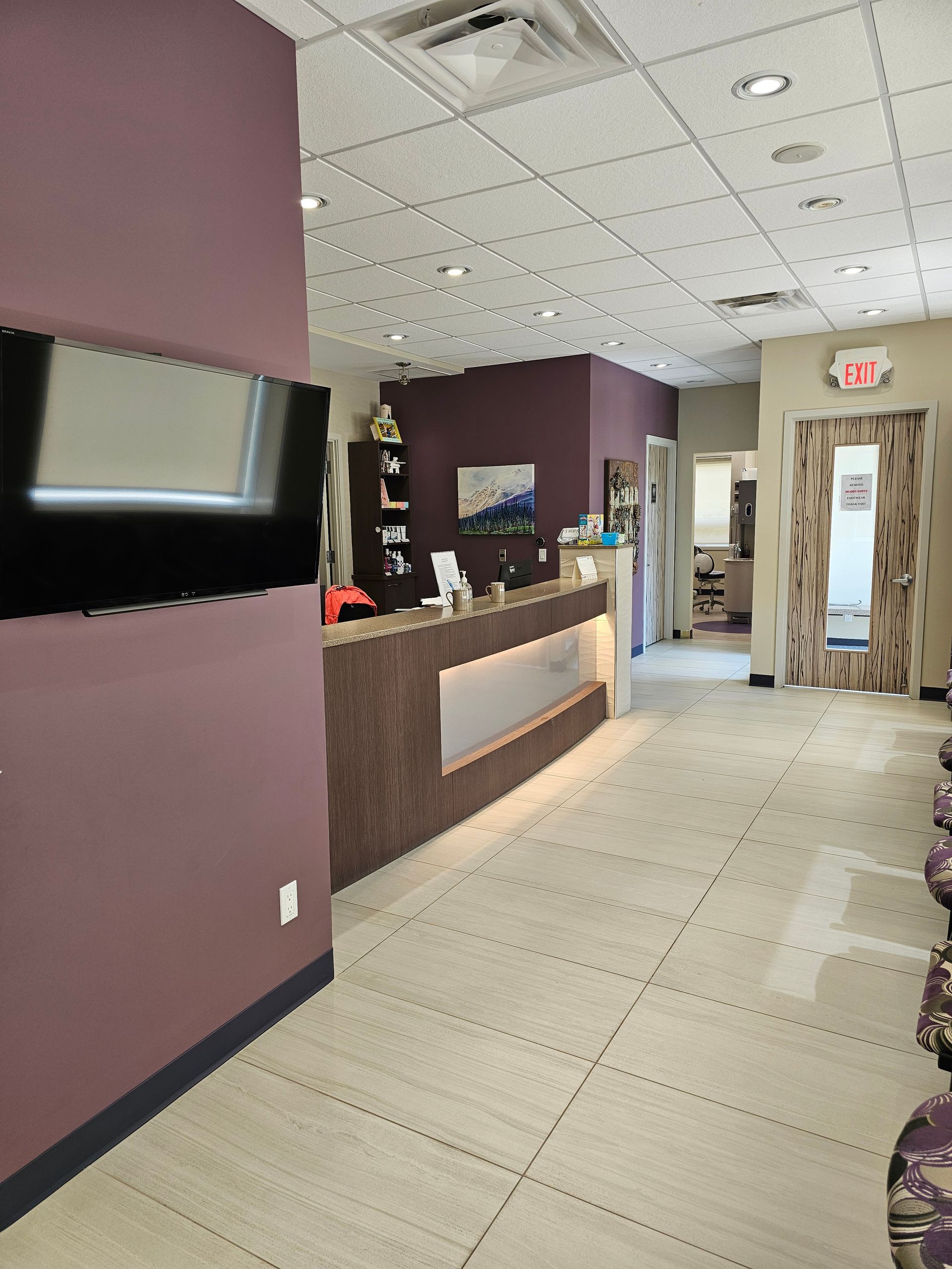 A modern office reception area with a dark wood desk, purple wall, wall-mounted television, and beige tiled flooring.