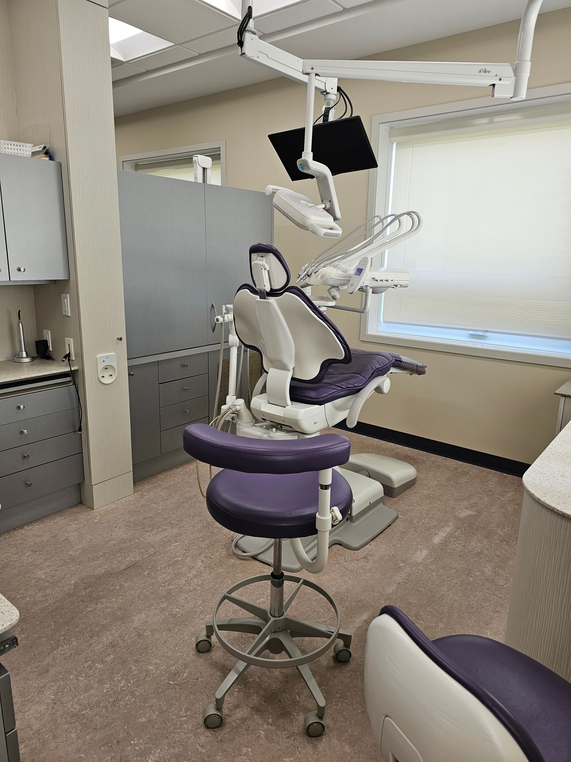 A professional dental operatory room featuring a purple dental chair, rolling stool, and equipment mounted on a ceiling arm.