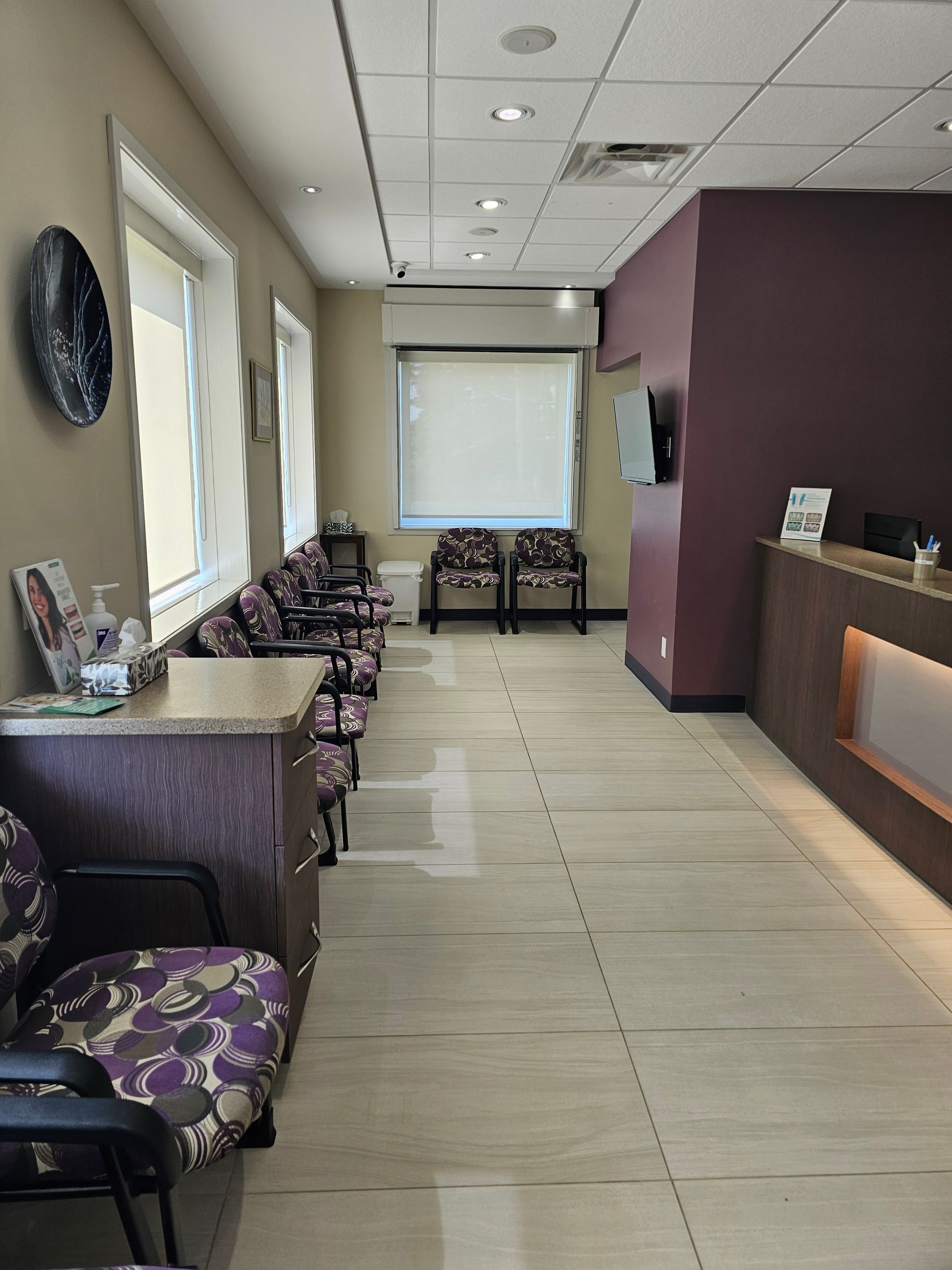 A waiting room with patterned chairs, a wooden reception desk, neutral walls, and a large window with white blinds.