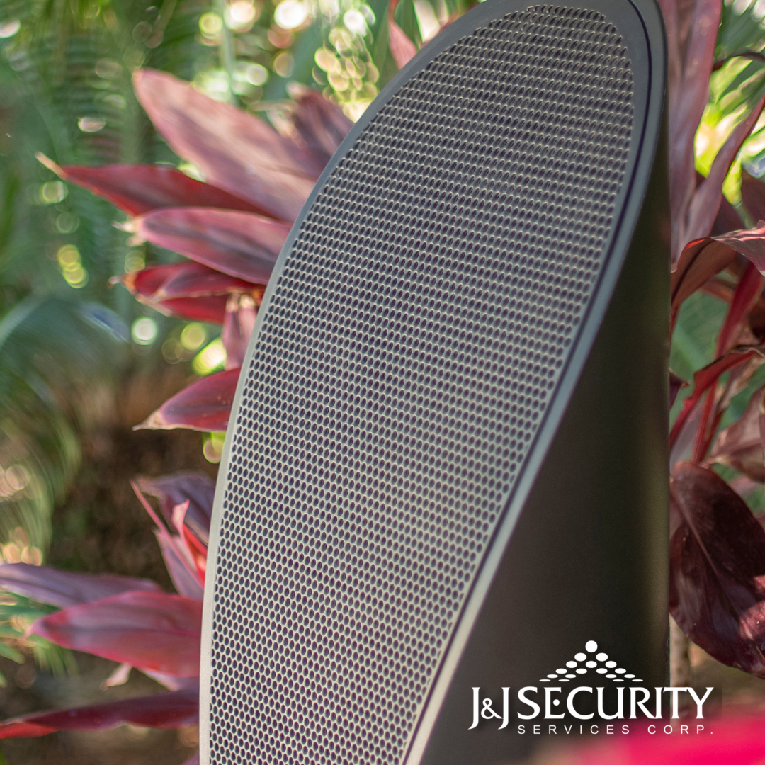 Close-up of a black outdoor speaker with a mesh grill, set amongst red plants.