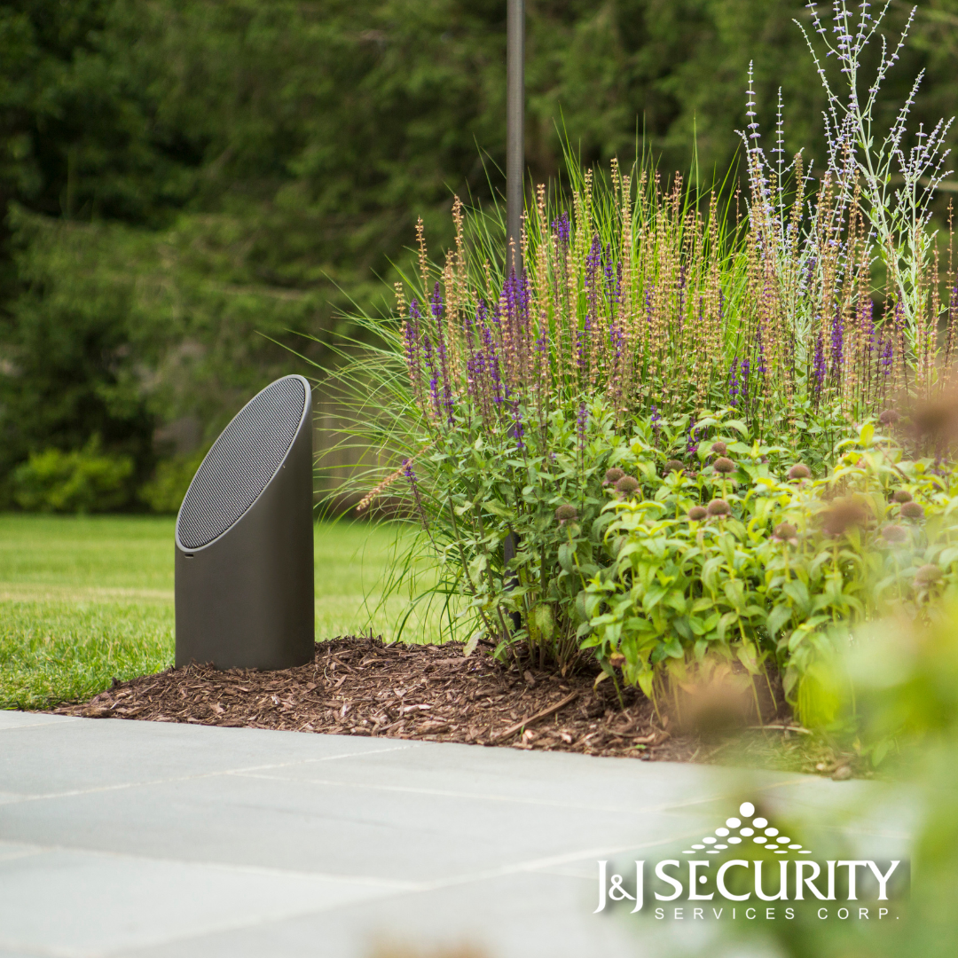 Dark gray outdoor speaker near a flower bed, on a stone path with green grass.