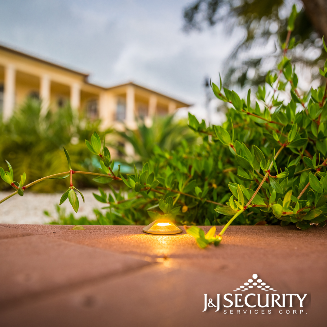 A low outdoor light illuminates a brick walkway with green foliage and a beige building in the background.