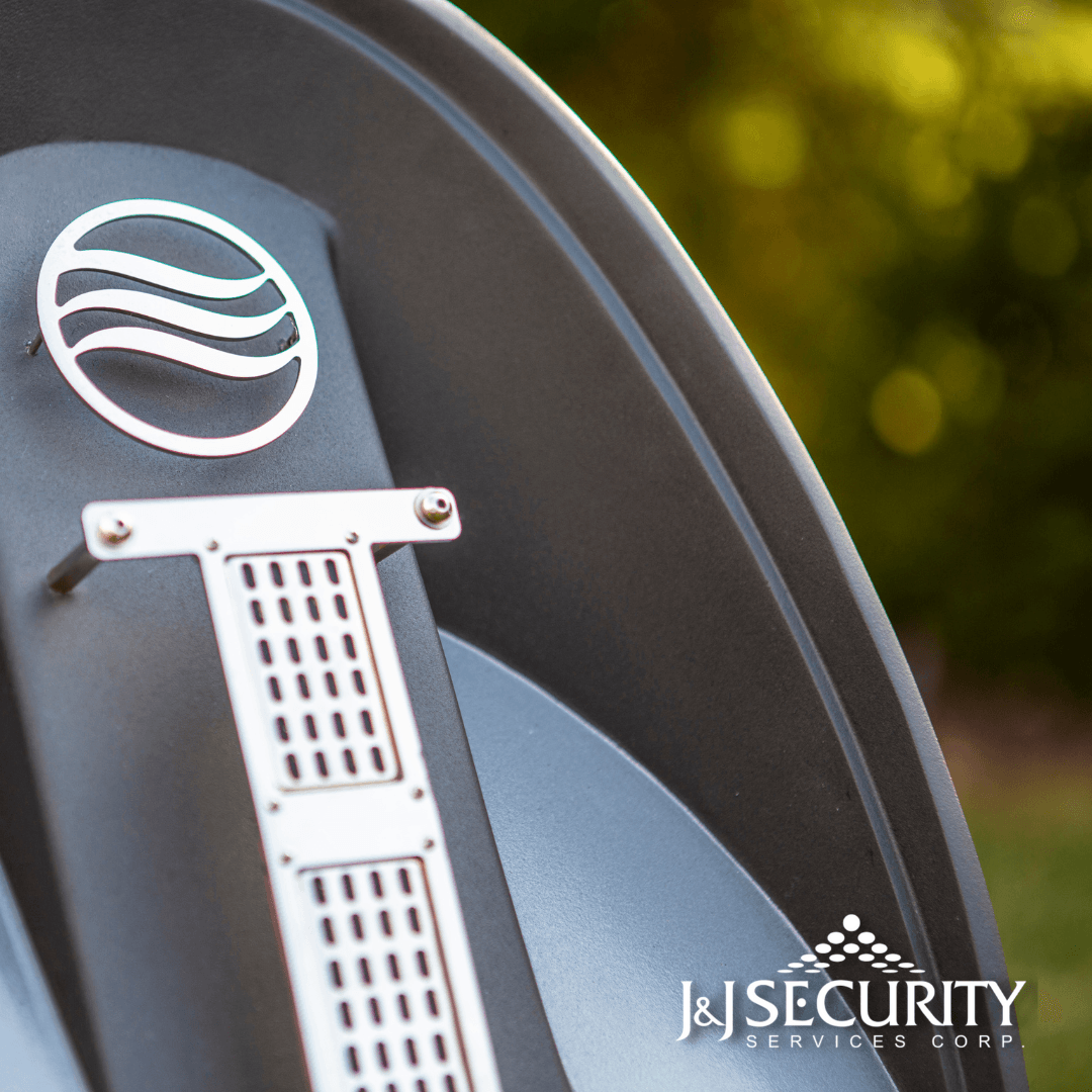 Close-up of a dark gray outdoor fixture with a silver logo and air vent. Blurred green foliage in the background.