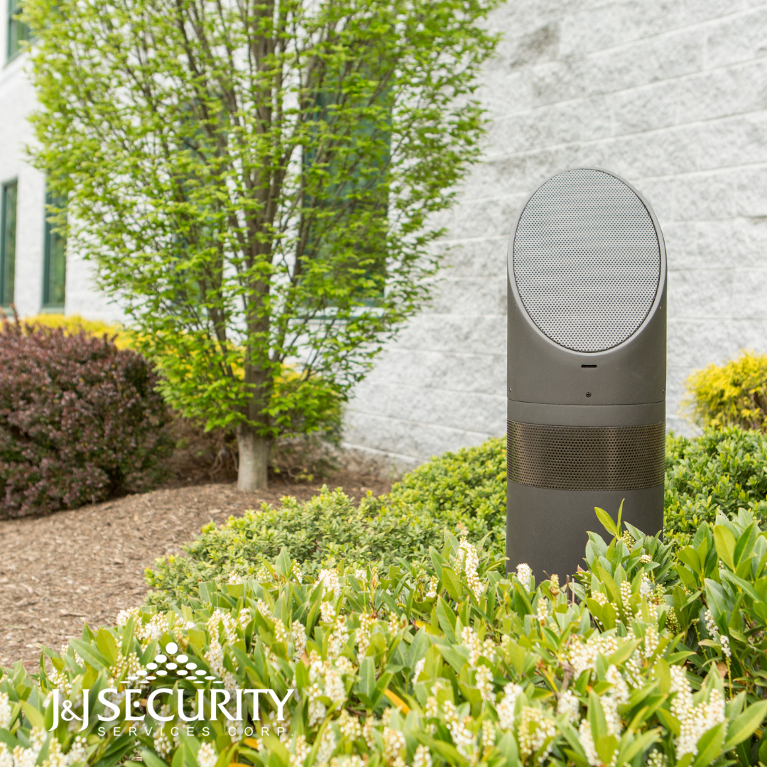 Security speaker in a garden setting, with plants and a building in the background.
