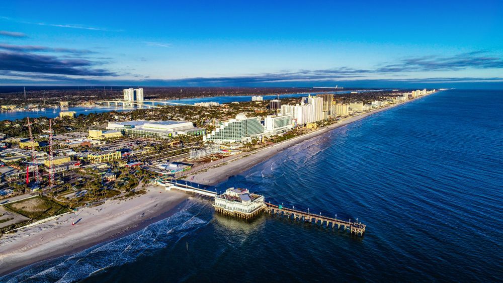 Aerial view of Daytona Beach, Florida, with pier, buildings, and ocean under a blue sky.
