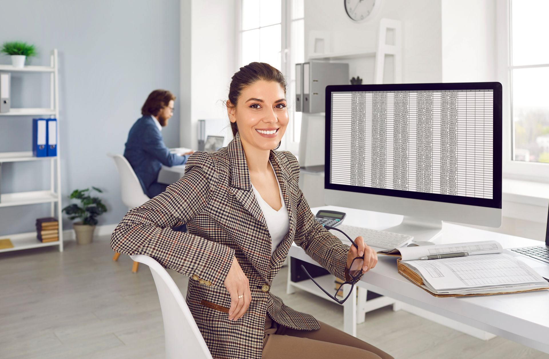 A woman sits at a desk with a computer displaying a spreadsheet, while a man works in the background