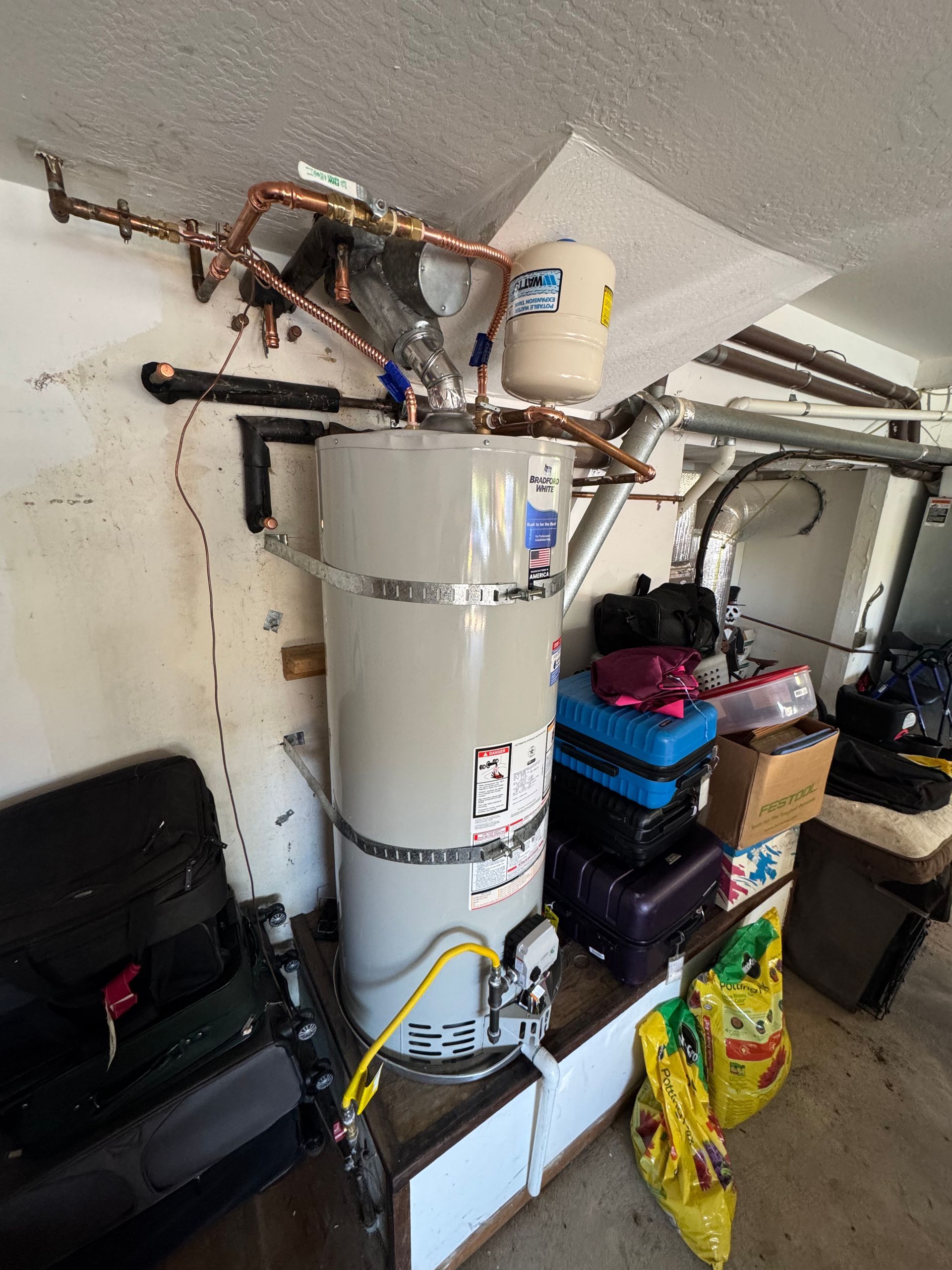 Water heater and plumbing in a garage storage area beside stacked boxes and bins.