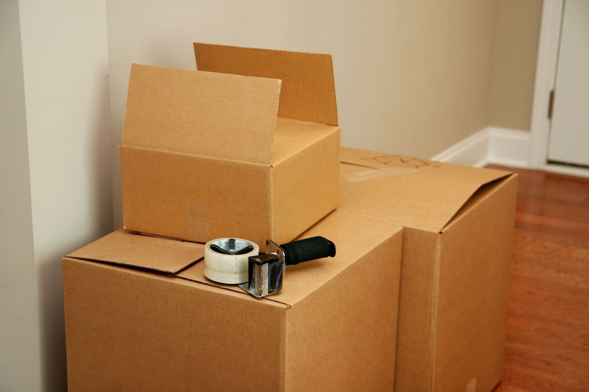 Cardboard moving boxes stacked in a corner, with packing tape and a cutter on top.