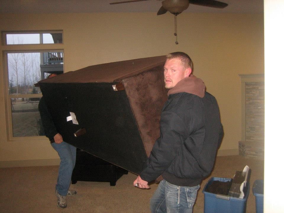 Two people carrying a large black couch through a sparsely furnished room