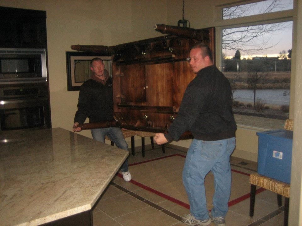 Two men carrying a wooden piano through a kitchen with a large island and window.