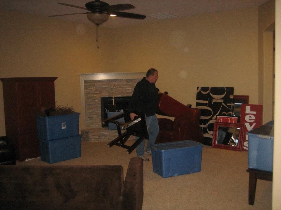Person moving furniture in a sparsely furnished room with blue storage bins and a ceiling fan.