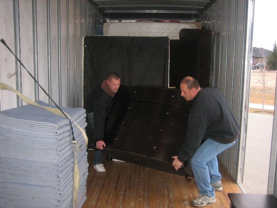 Two men loading a large black object into a truck trailer with folded mats stacked on the left.