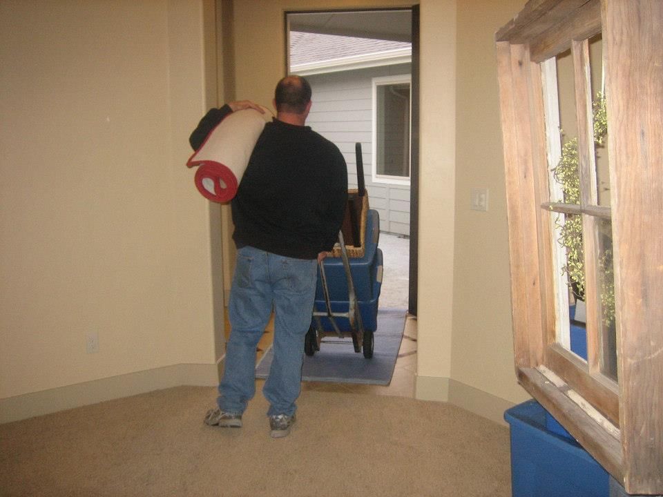 Man carrying a rolled carpet through a doorway in a room with carpet and windows