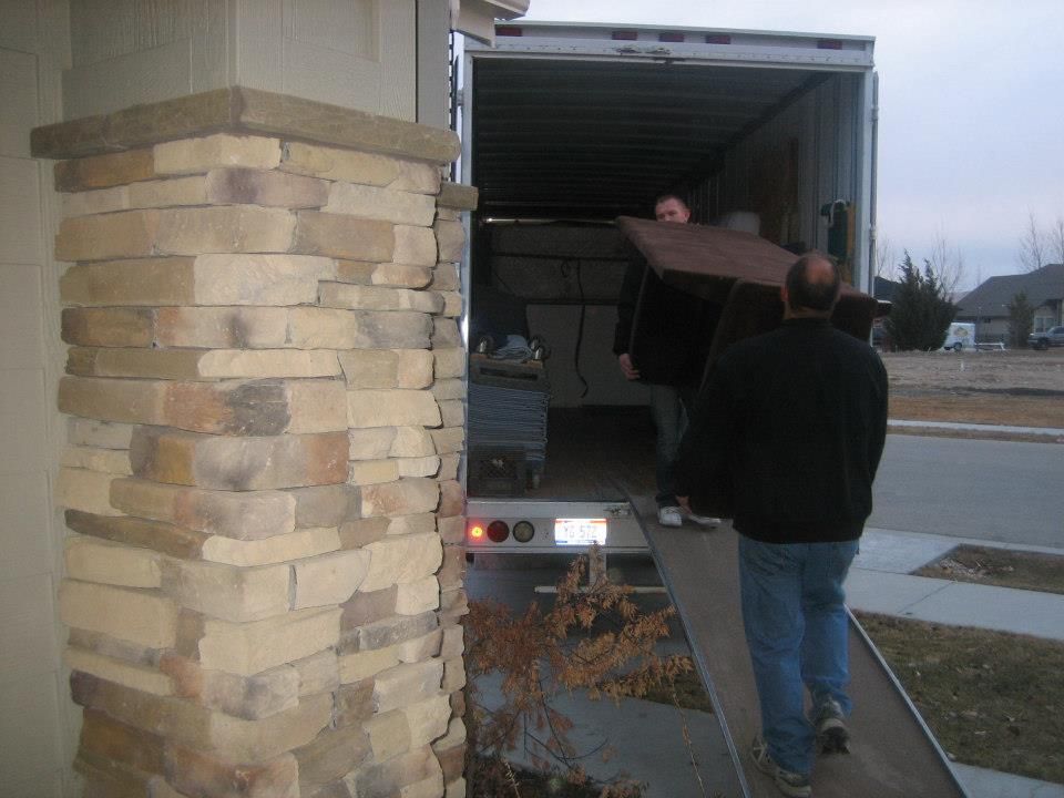 Two people unloading a large box from a truck at a driveway beside a stone pillar.