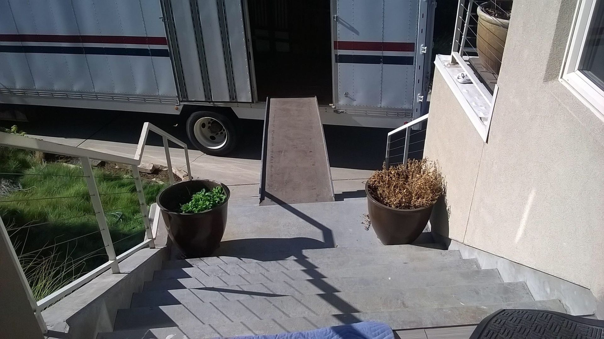 Outdoor steps beside a white building, with potted plants and a ramp leading to a parked trailer.