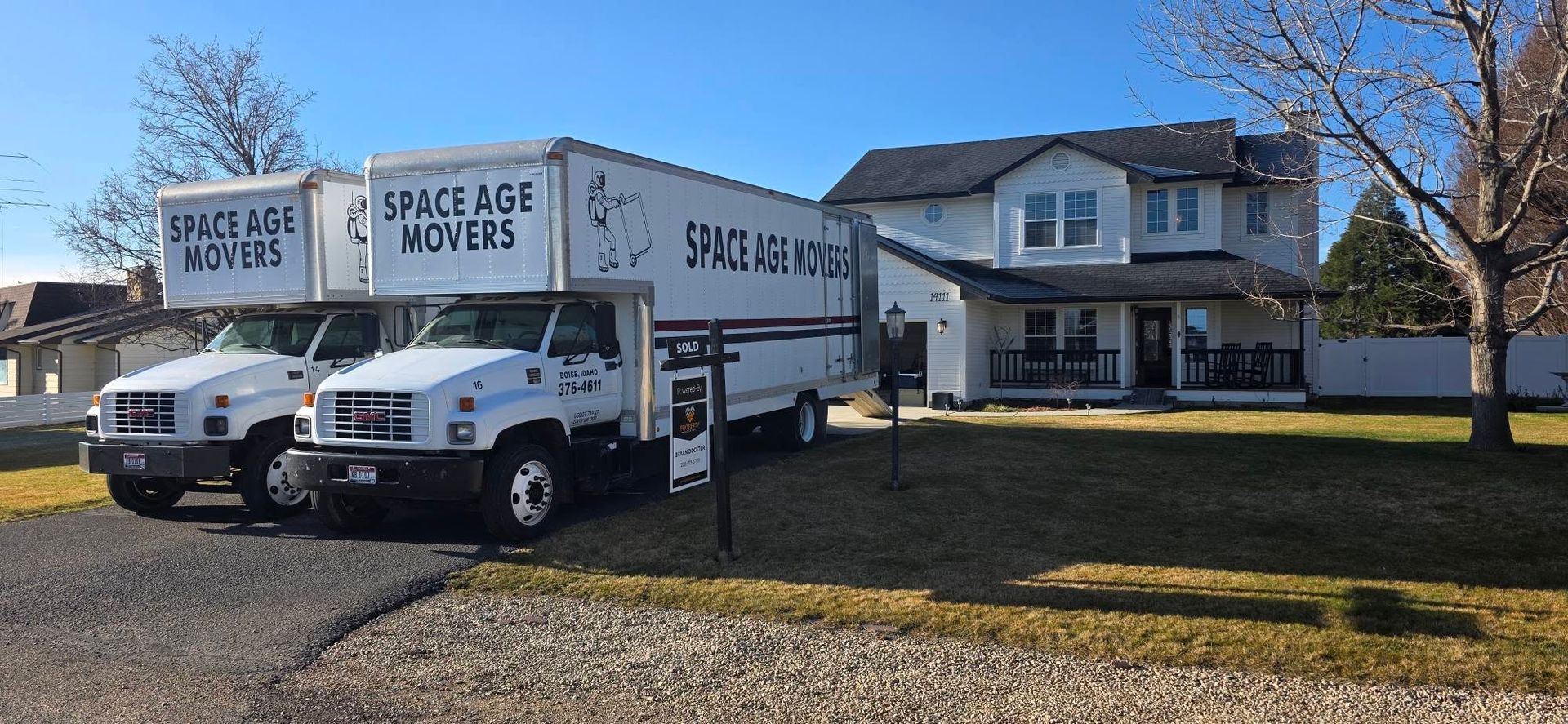 Two white moving trucks parked on a lawn in front of a house on a sunny day.