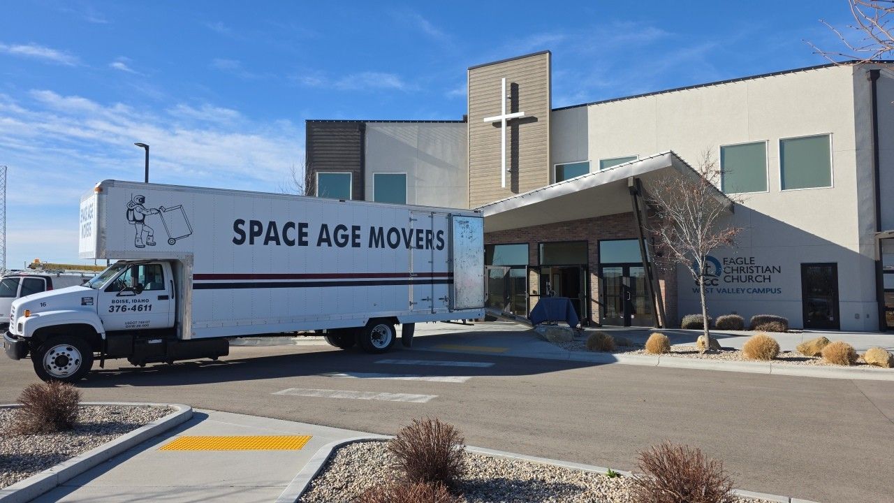 Moving truck labeled “SPACE AGE MOVERS” parked outside a modern building in bright sunlight