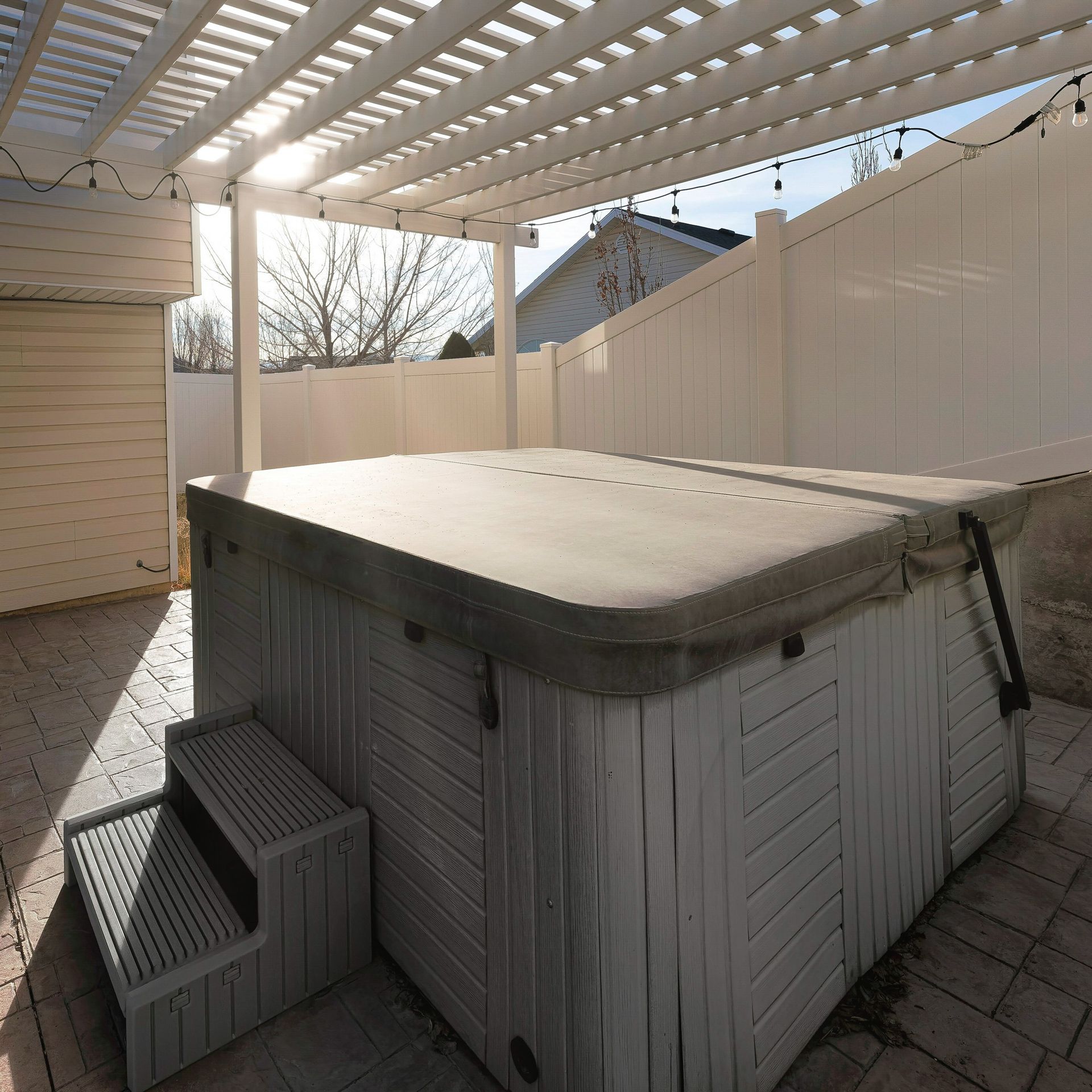 Covered backyard hot tub under a white pergola beside a fence on a patio.