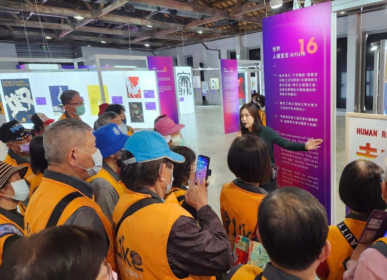 Group of people listening to a guide in a gallery. The guide points to a purple panel. People wear orange vests.