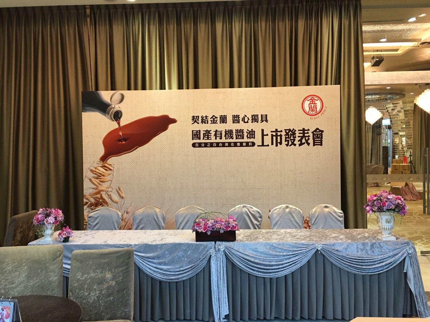 Press conference setup with banner, table, chairs, flowers, and draped curtains.