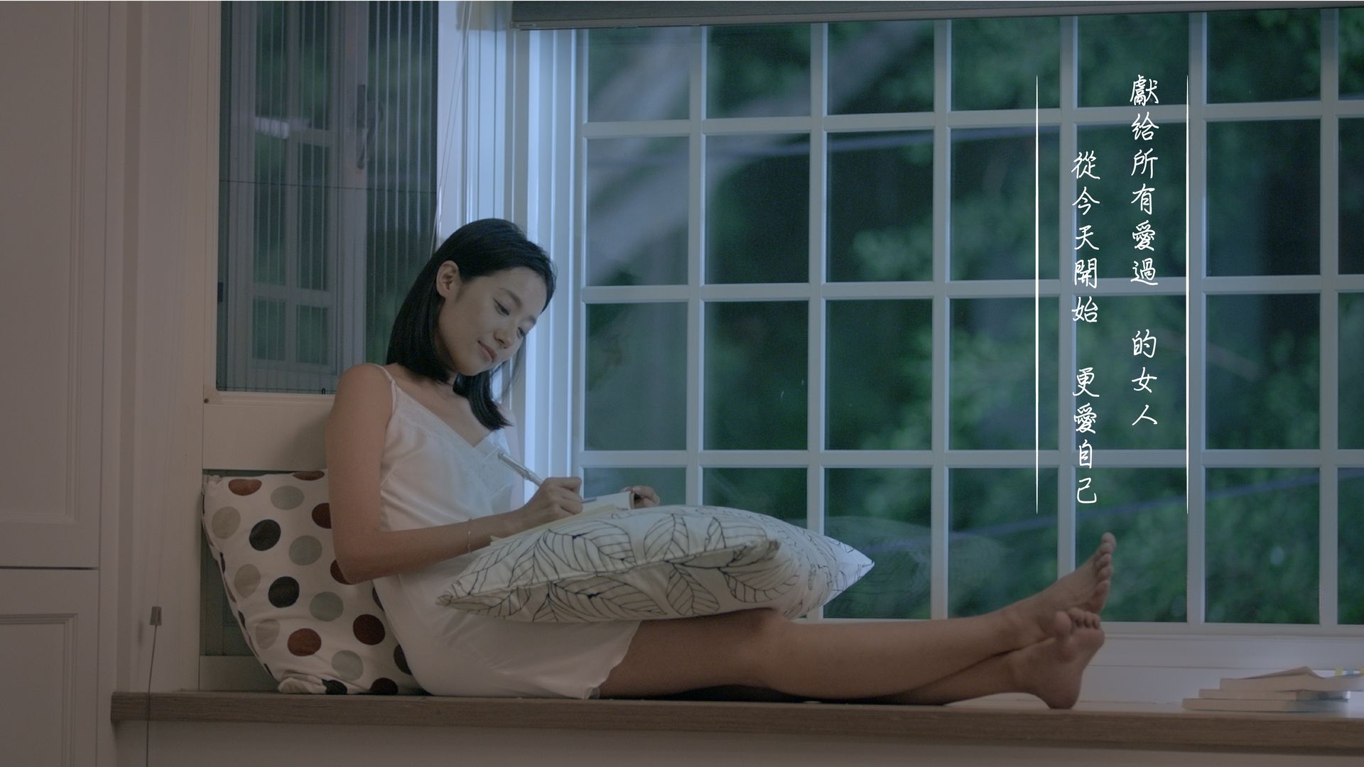 Woman sits in window, writing on paper, legs extended. Gray light, pillow, and outdoor foliage visible.
