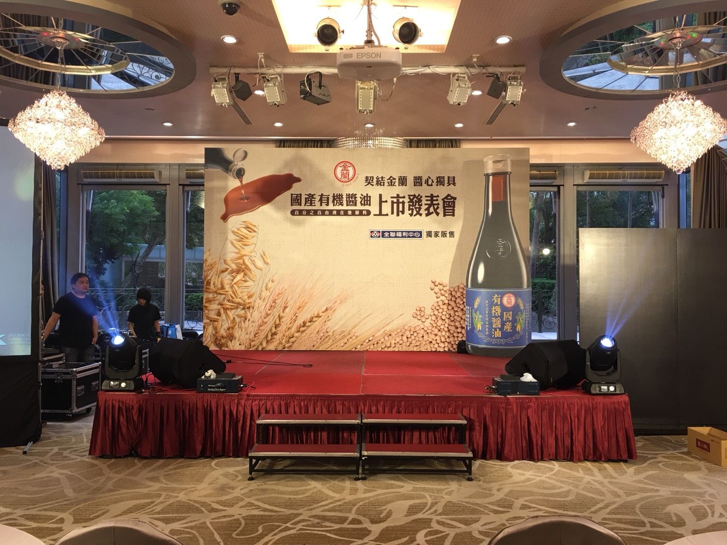 Stage set up for an event, featuring a backdrop with a bottle illustration. Red stage, lighting, and a few people.