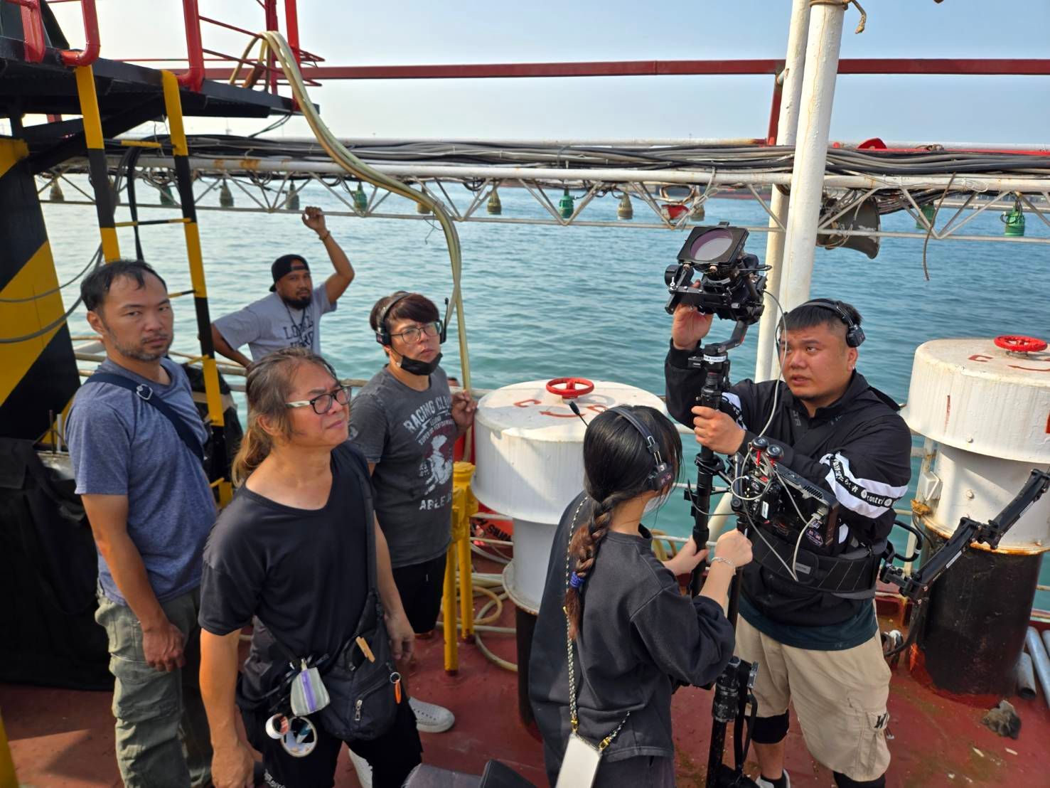 Film crew on a boat filming. A man holds a camera, others wear headsets, watching the ocean.