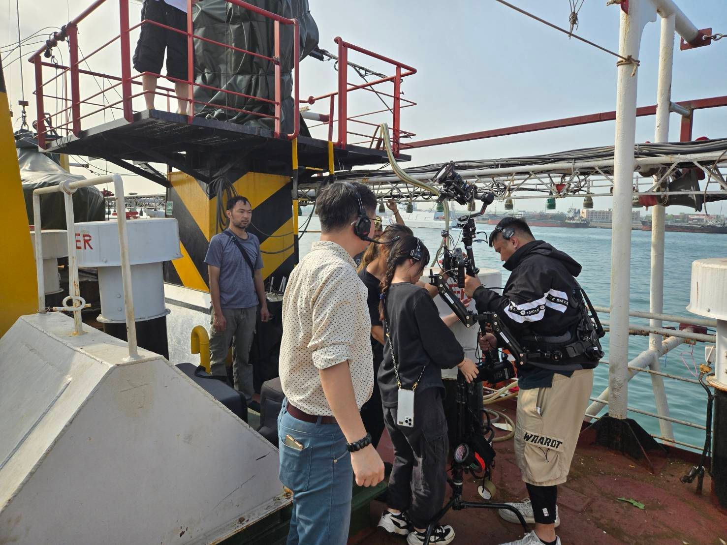 People on a boat deck, rigging equipment. Some wear headphones, working near water.