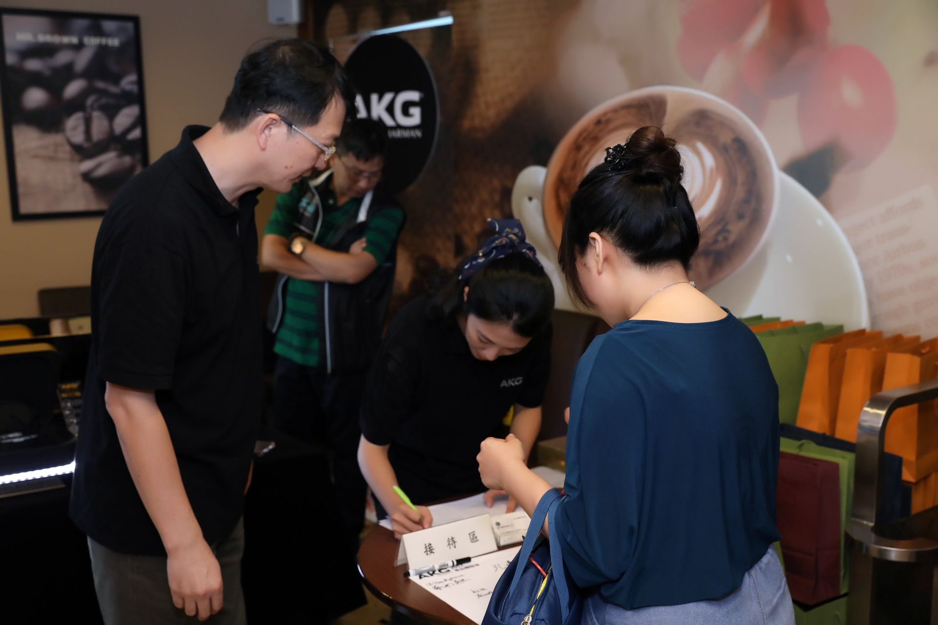 People at a coffee shop counter: customer signing, employee assisting, another person observing, coffee-themed background.
