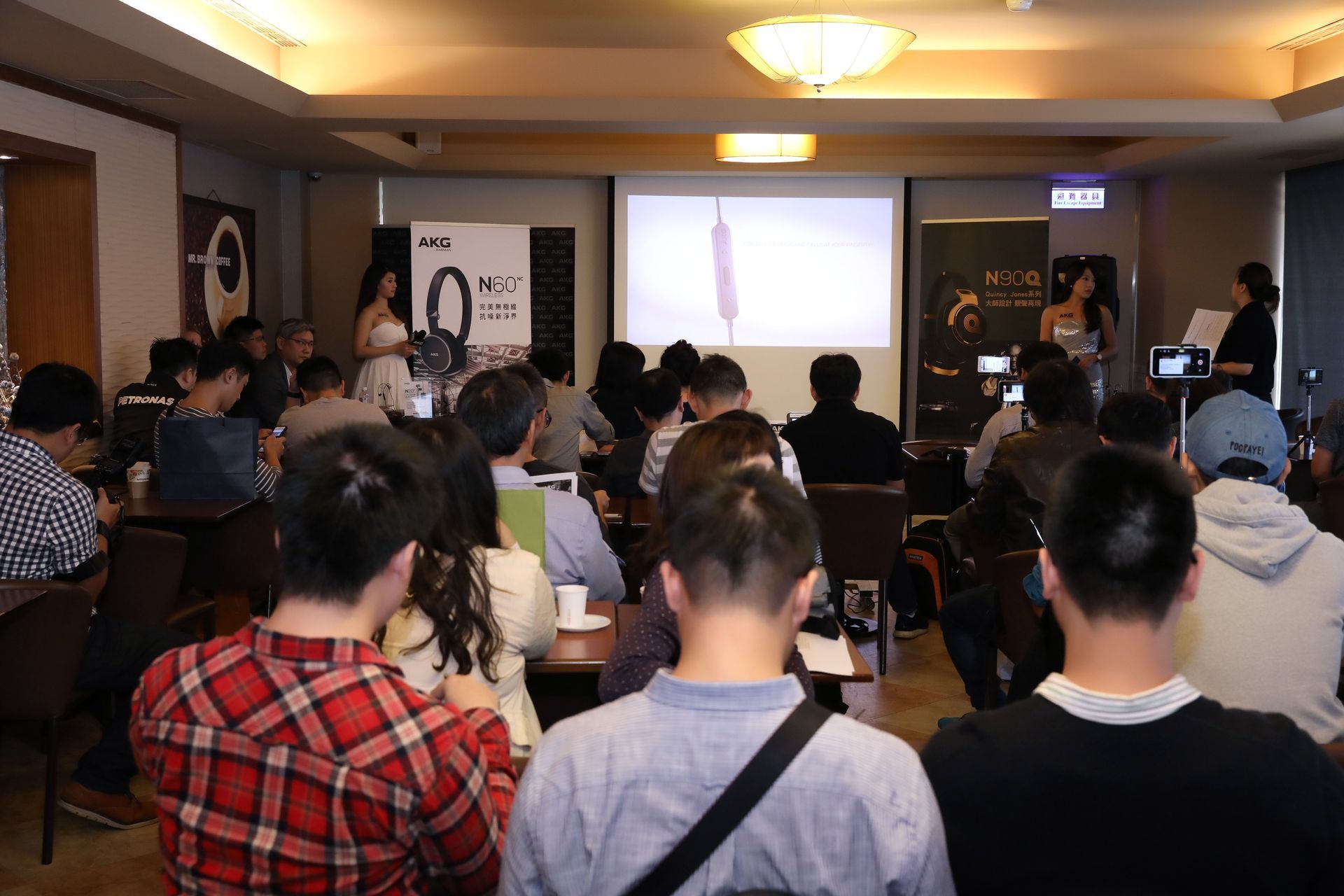Audience in a presentation room watches a screen. Two presenters stand nearby.