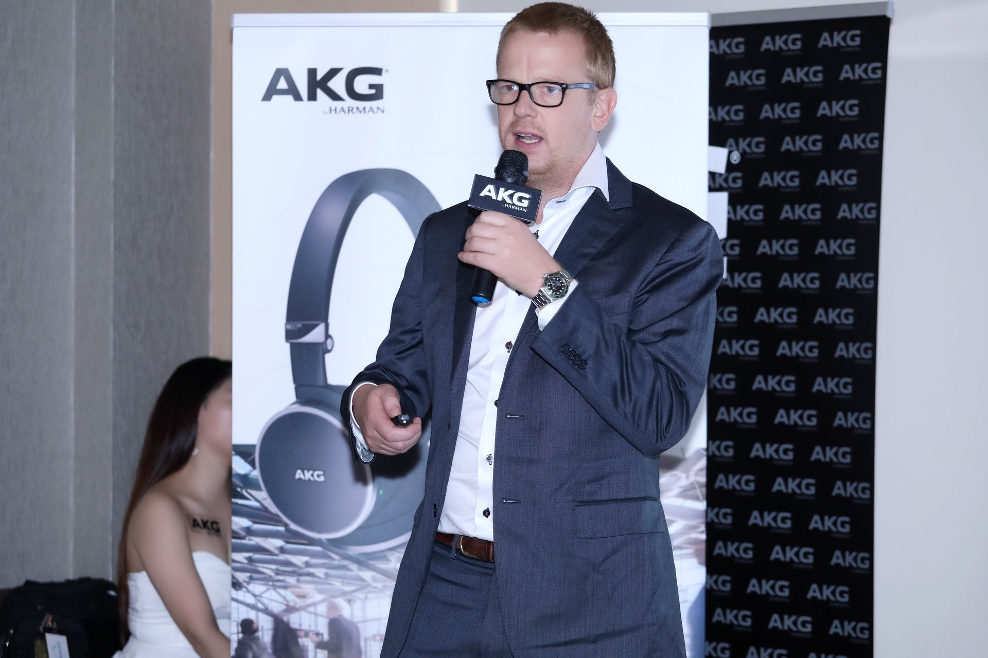 Man in suit speaking at an AKG headphones product launch event. AKG logo and headphones in background.