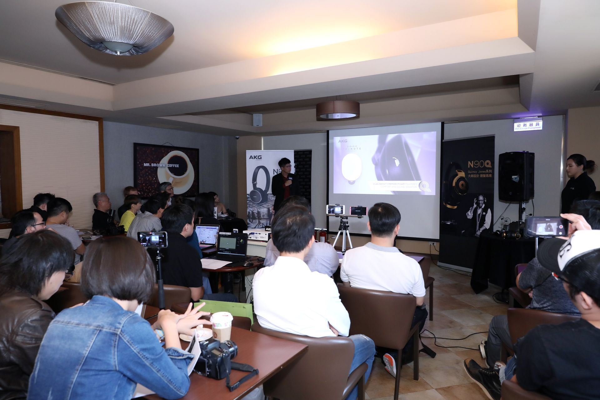 People attending a presentation in a room with a screen; presenters are visible.