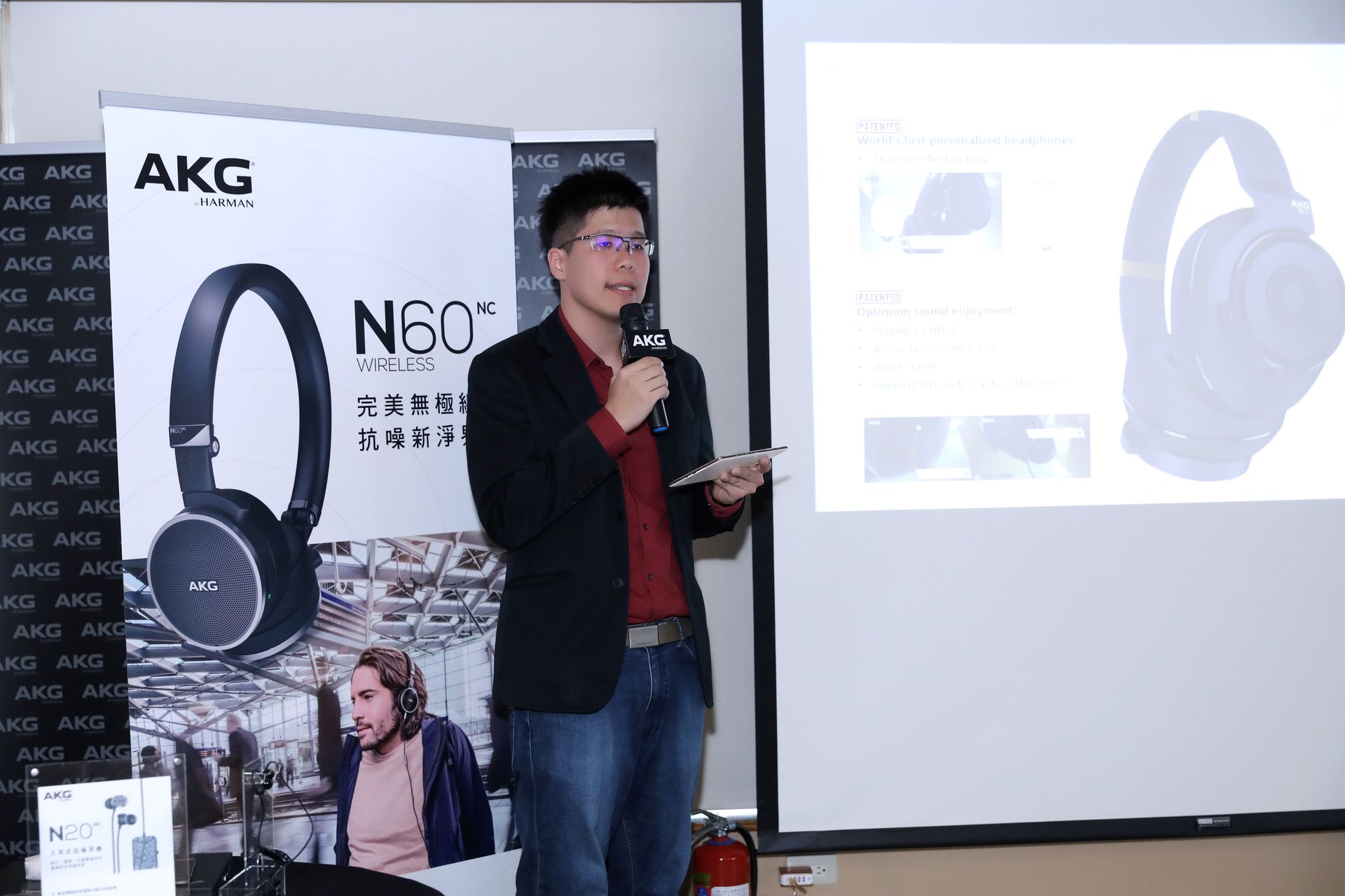Man presenting AKG N60NC headphones at event, holding a microphone. Poster and screen show product details.