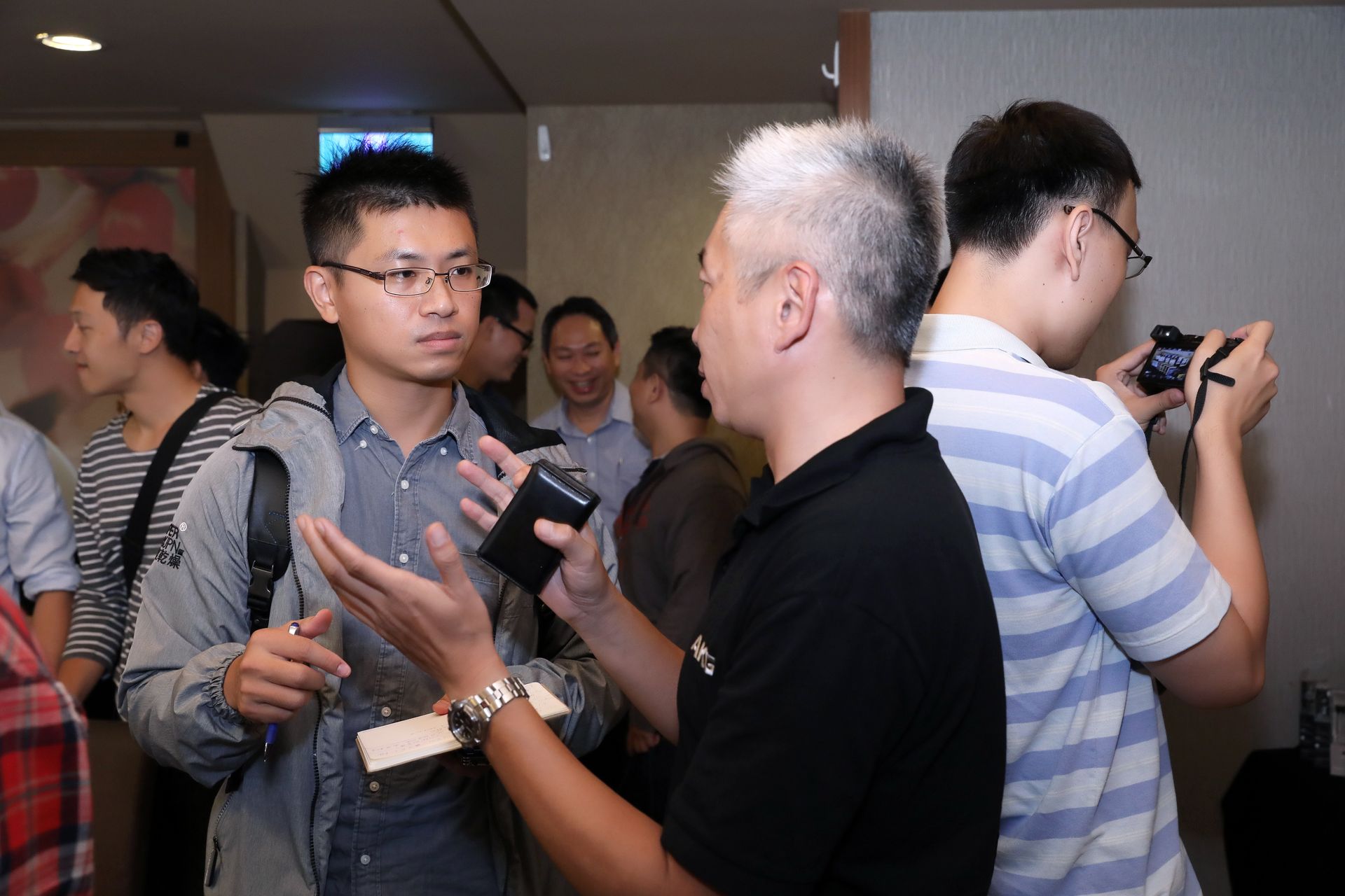 Two men talking, one holding a device. Several other people in background, some looking at them. Indoors.
