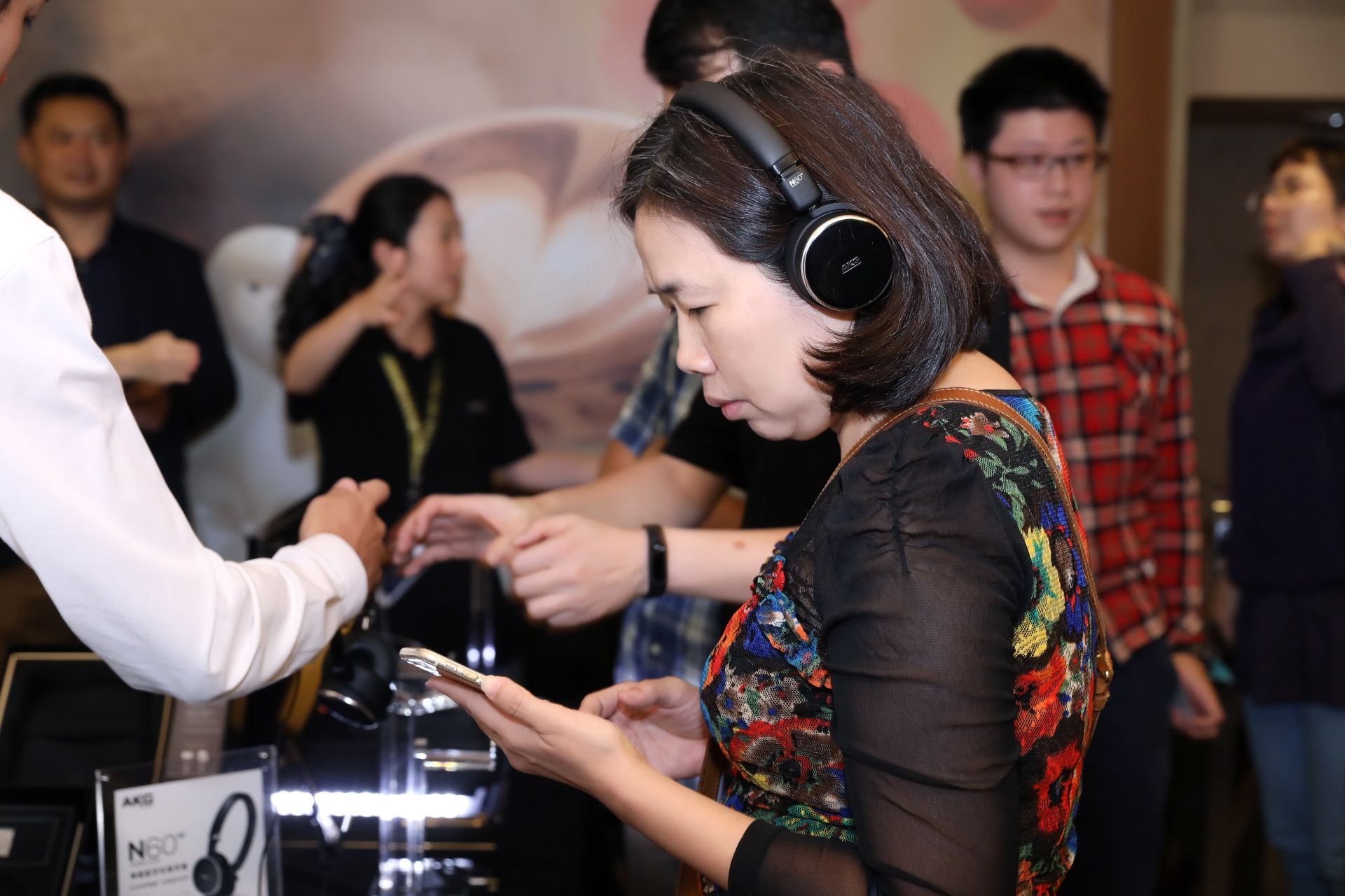 Woman wearing headphones, using a phone. People around a display table at a tech event.